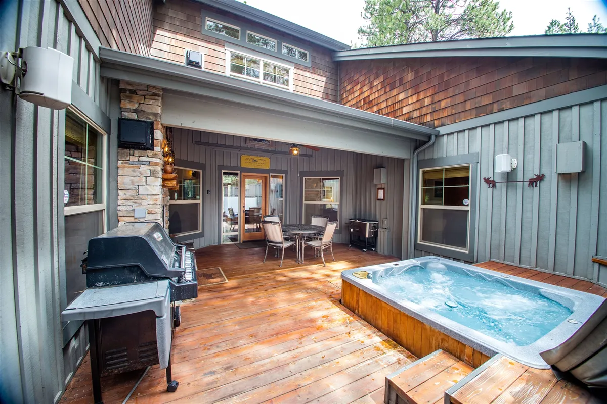 An exterior view of the deck featuring a hot tub, dining table, and grill. The hot tub is positioned to catch sunset views, while the dining area invites guests to enjoy meals outdoors. The space is open yet private, surrounded by natural elements.