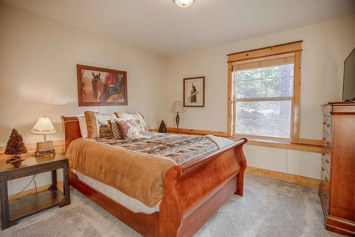 A warm, spacious bedroom with a classic wooden bed frame, decorative pillows, artwork above the bed, and a nightstand with a glowing lamp. The setting is restful and welcoming, designed for peaceful nights and quiet mornings.