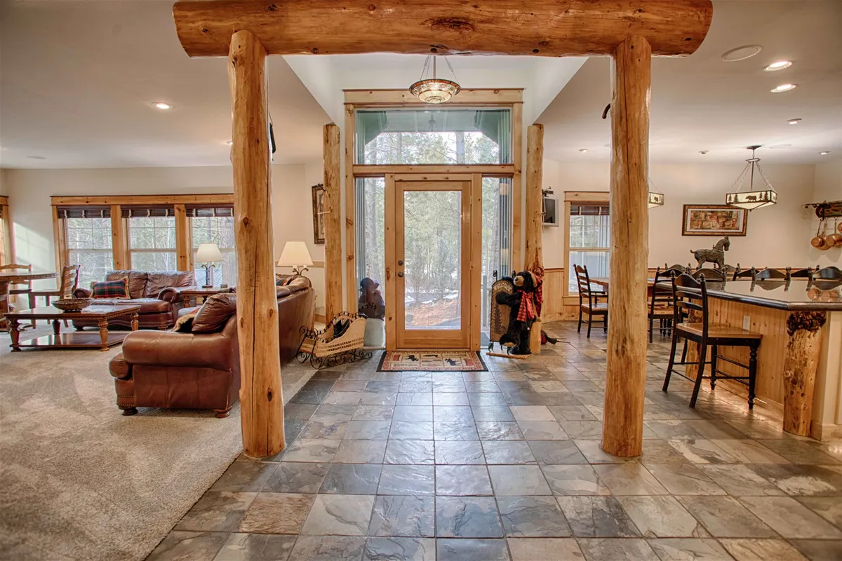 A welcoming open-plan living and dining area with log pillars, a front door leading to the outdoors, and cozy furnishings. The space features a dining table set for gatherings, a fireplace, and large windows that let in natural light and showcase snow falling outside.