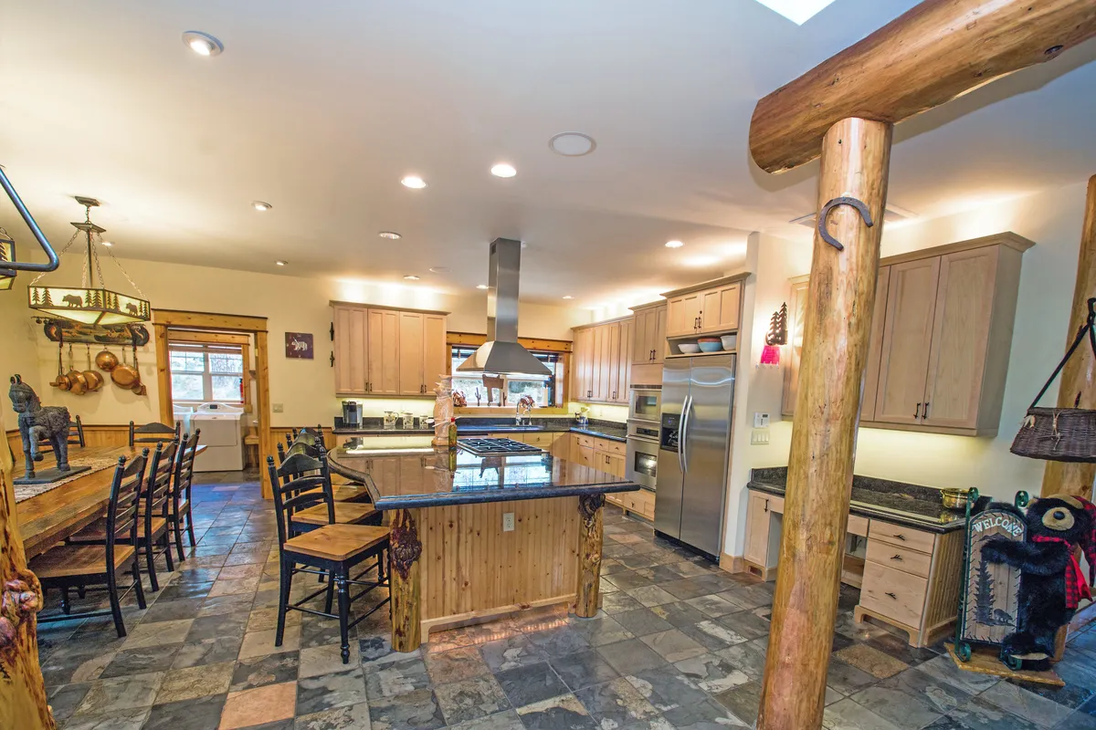 A view of the expansive chef's kitchen, complete with a large central island, granite countertops, stainless steel appliances, and exposed wooden beams. The kitchen opens directly to a spacious dining area, illustrating the open-concept design ideal for cooking together and group meals.