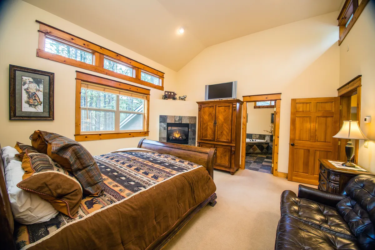 A bedroom with a decorative bed, wooden furniture, large windows with nature views, and a fireplace. The space is warm and comfortable, ideal for unwinding, reading, or catching up on work in a relaxing environment.