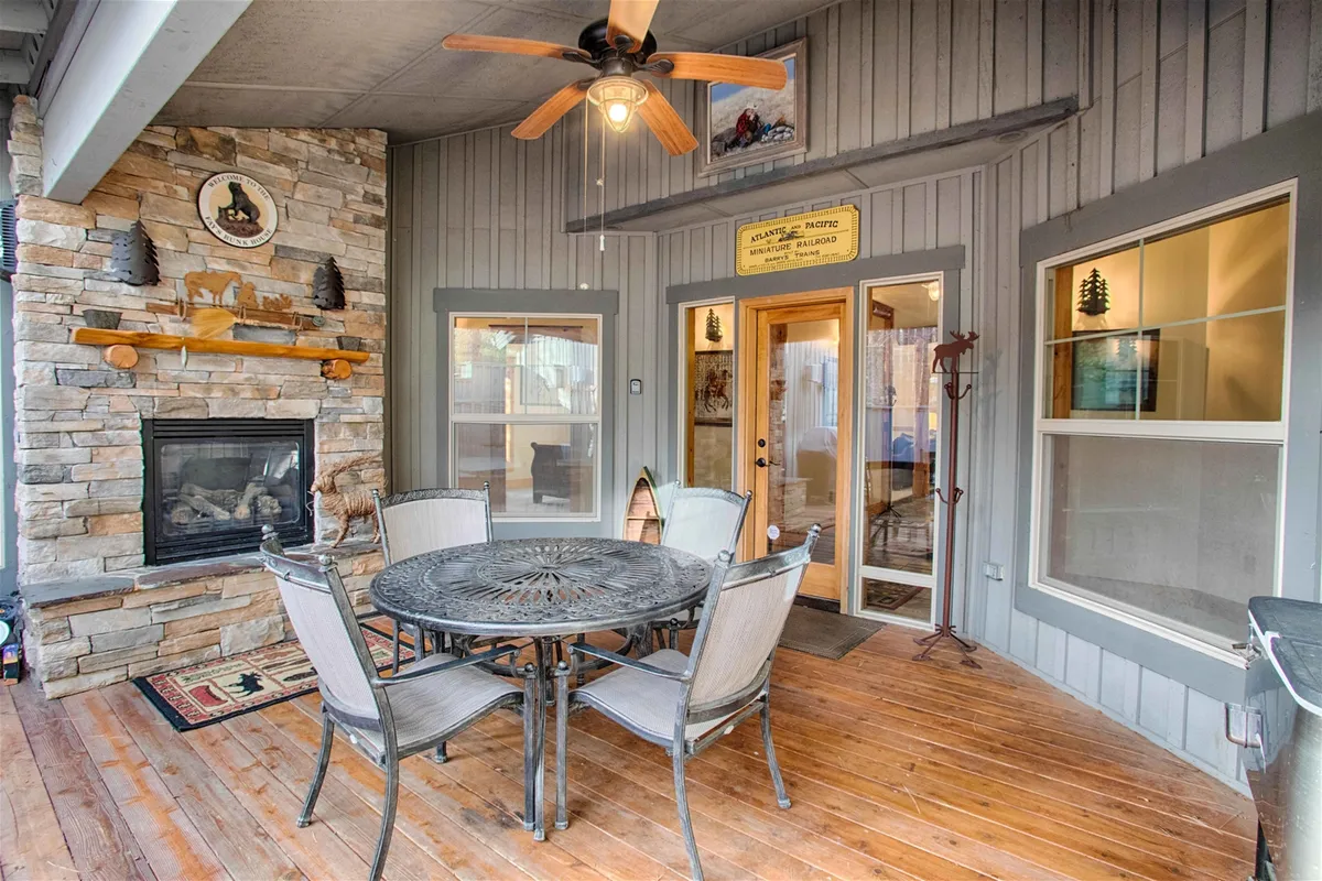 A covered outdoor deck with a stone fireplace as a central focal point. The area also features a dining table set for meals, a ceiling fan overhead for comfort, and decorative elements on the privacy wall. The setting is cozy and sheltered, with warm lighting and plenty of space for gatherings.