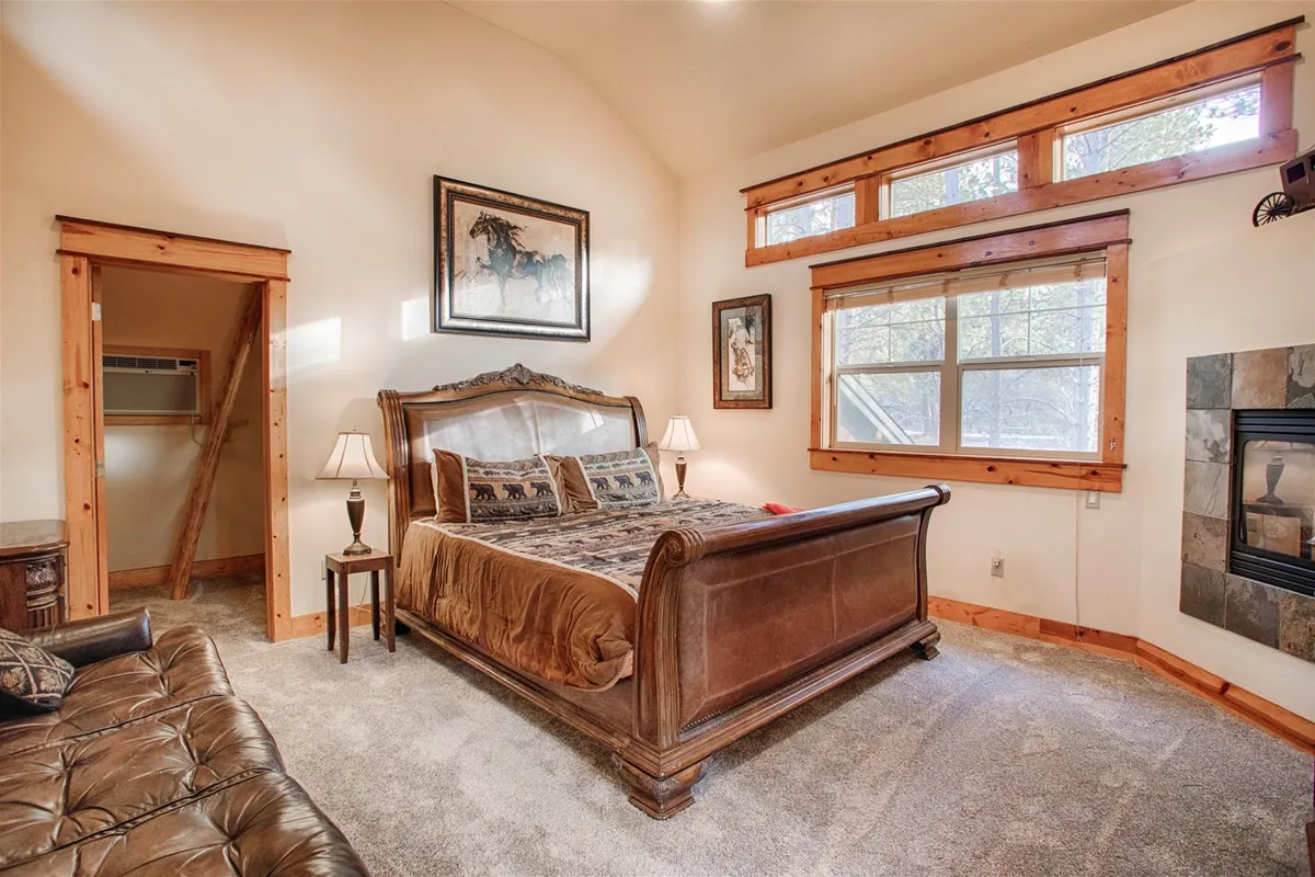 A spacious bedroom featuring a large wooden bed, an inviting fireplace, and wood-trimmed windows that fill the space with natural light. Artwork on the walls and comfortable bedding enhance the cozy atmosphere, suggesting both rest and relaxation in a serene setting.