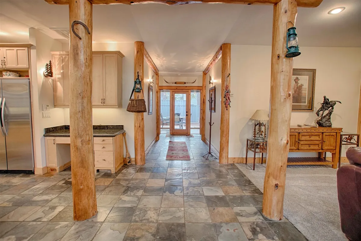 A wide hallway framed by wooden pillars and slate flooring, leading toward double doors. Decorative wall art and a side table with a lamp create a welcoming, communal feel ideal for gathering and social activities.